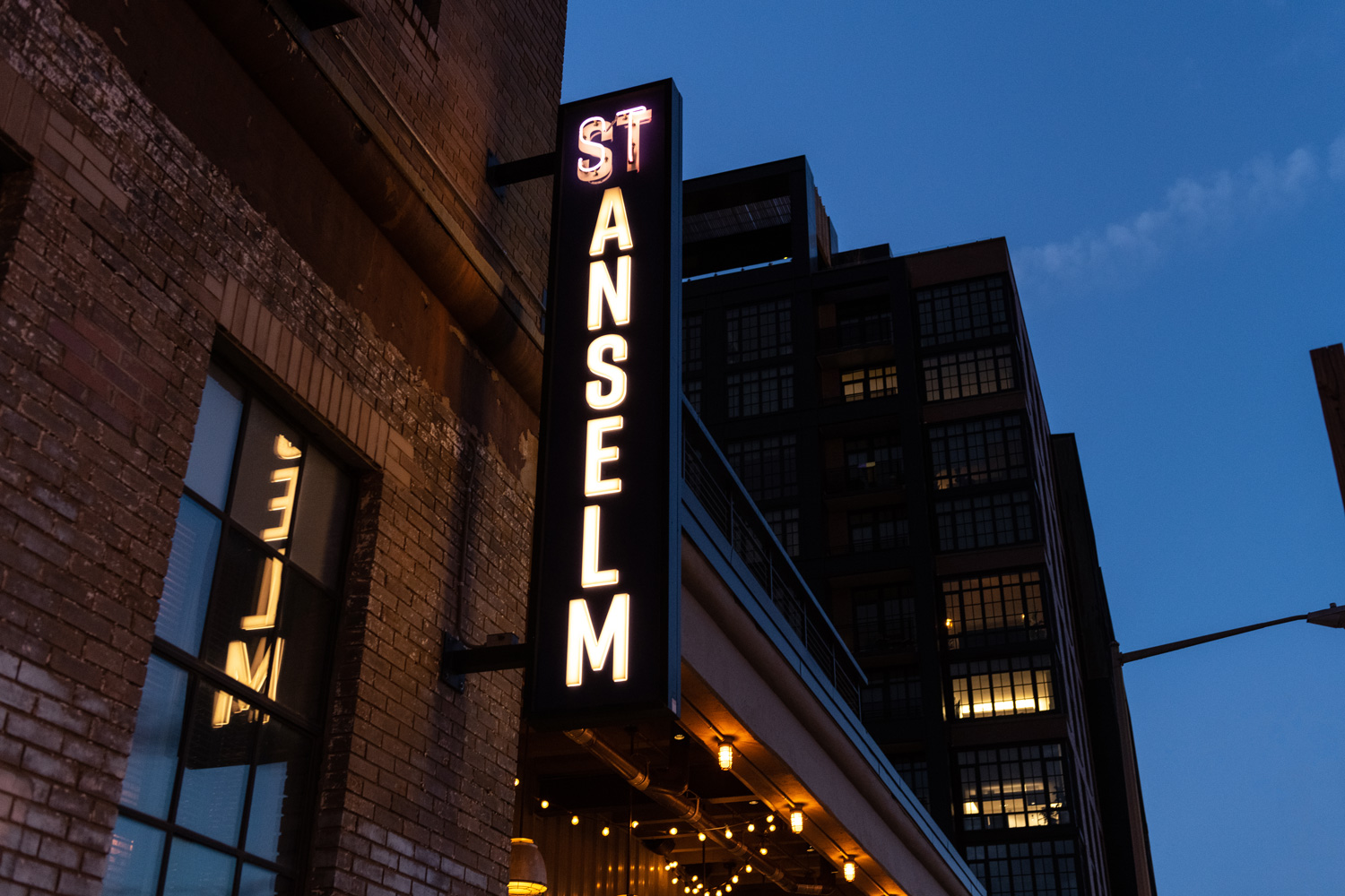Sidewalk view of the St. Anslem lit bar sign and city nightlife